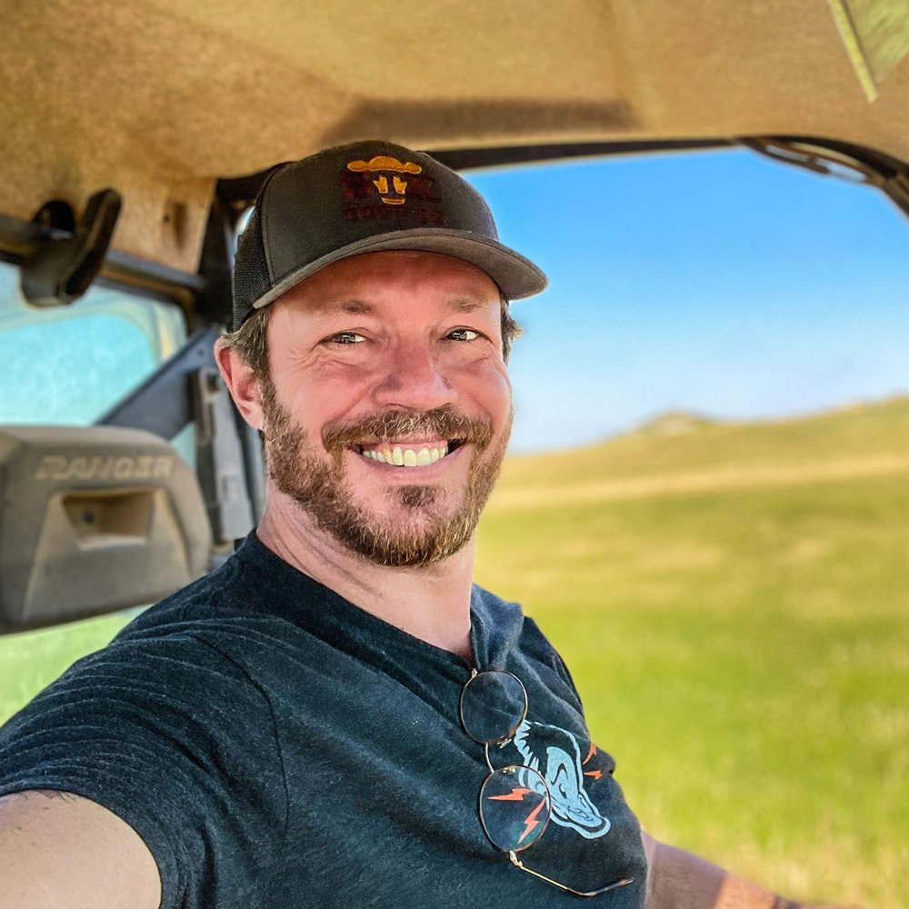 Headshot of Parable Pictures team member Dan Brooks smiling, wearing a ball cap and sitting inside a utility terrain vehicle in a sunny, open pasture