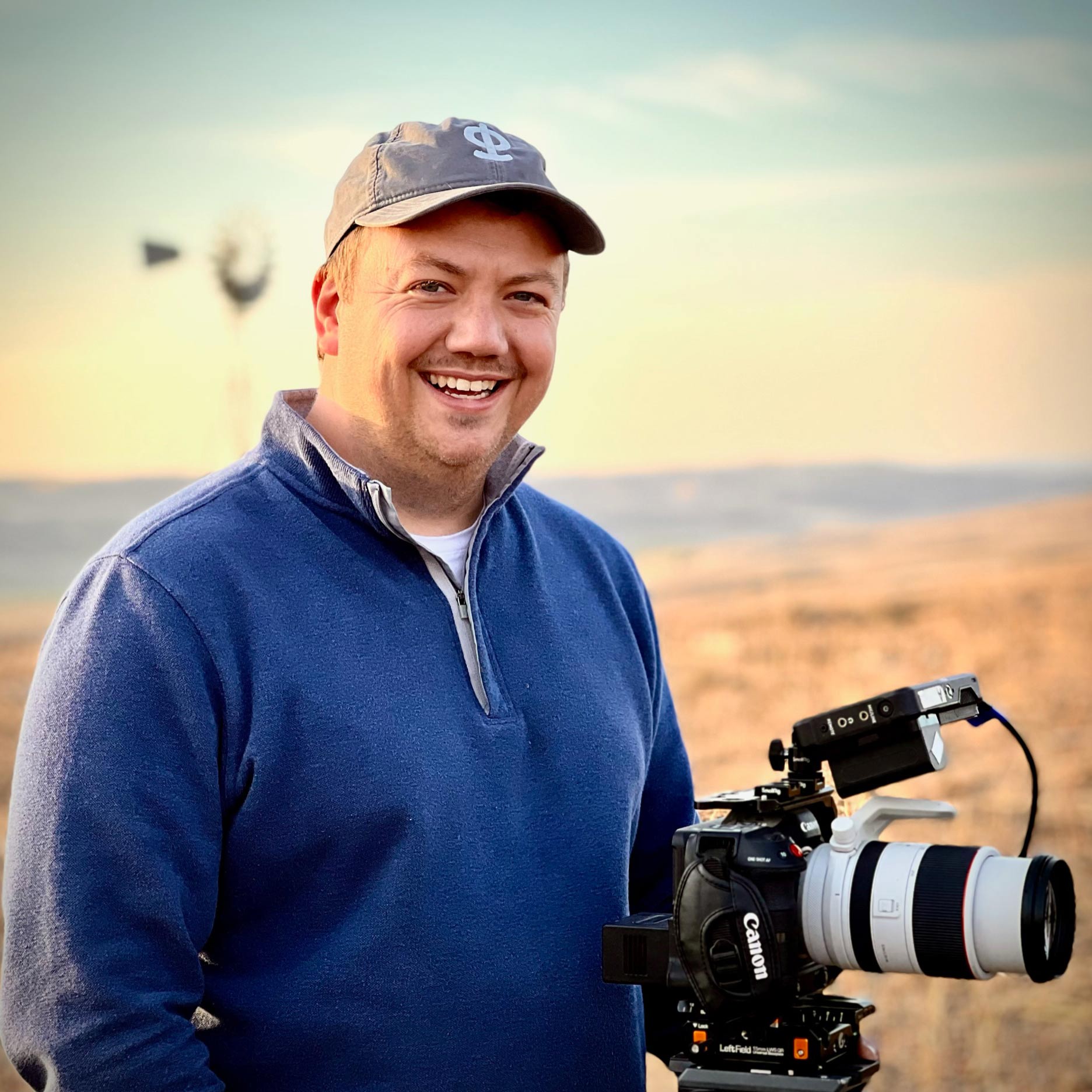 Headshot of Parable Pictures team member Will Brooks smiling, wearing a ball cap and standing with his Canon camera in an open field on a set